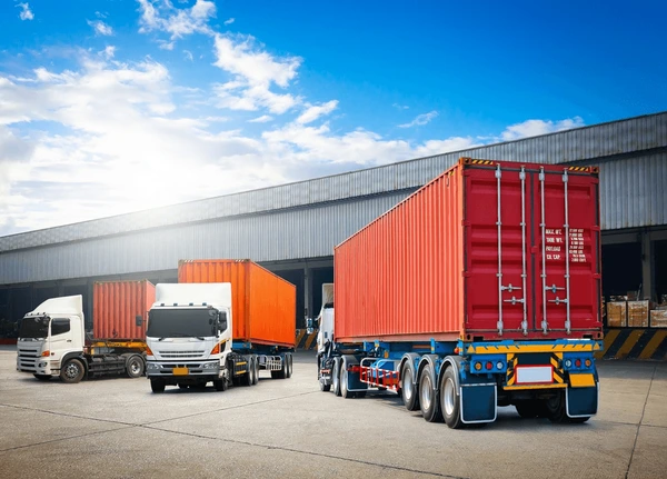 Multiple trucks with large orange shipping containers parked at a warehouse loading dock under a sunny sky. Priority Auto Shipping | Transportvibe.
