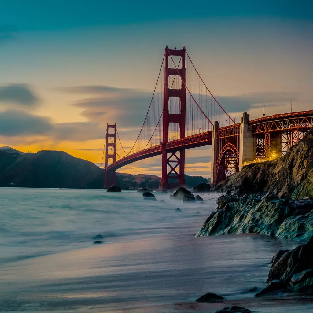 A stunning view of the Golden Gate Bridge in California , San Francisco at sunset, with waves gently hitting the rocky shoreline and a dramatic sky in the background. Transportvibe