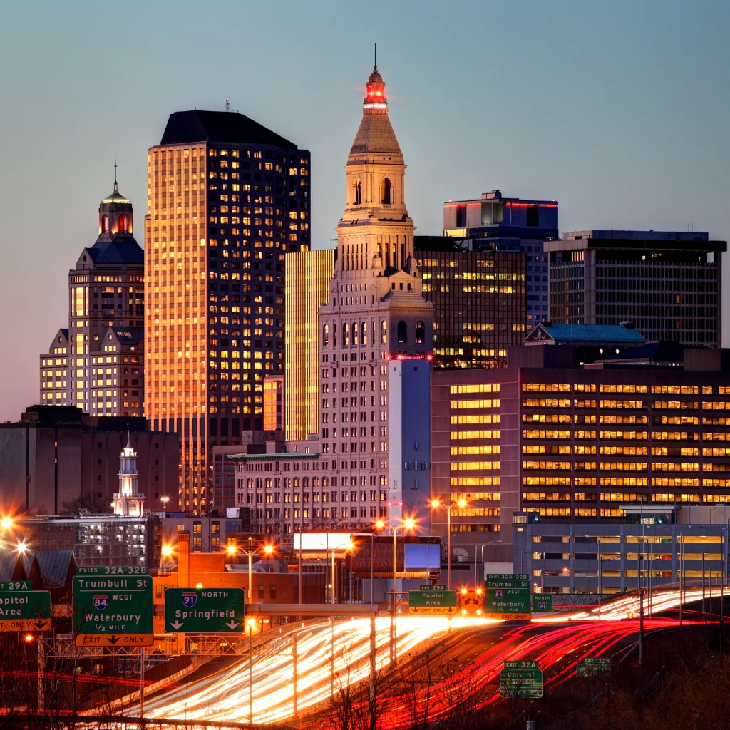 A breathtaking nighttime view of Hartford, Connecticut’s skyline, showcasing illuminated skyscrapers and historic architecture. Light trails from highway traffic add a sense of motion to the vibrant urban scene. Transportvibe