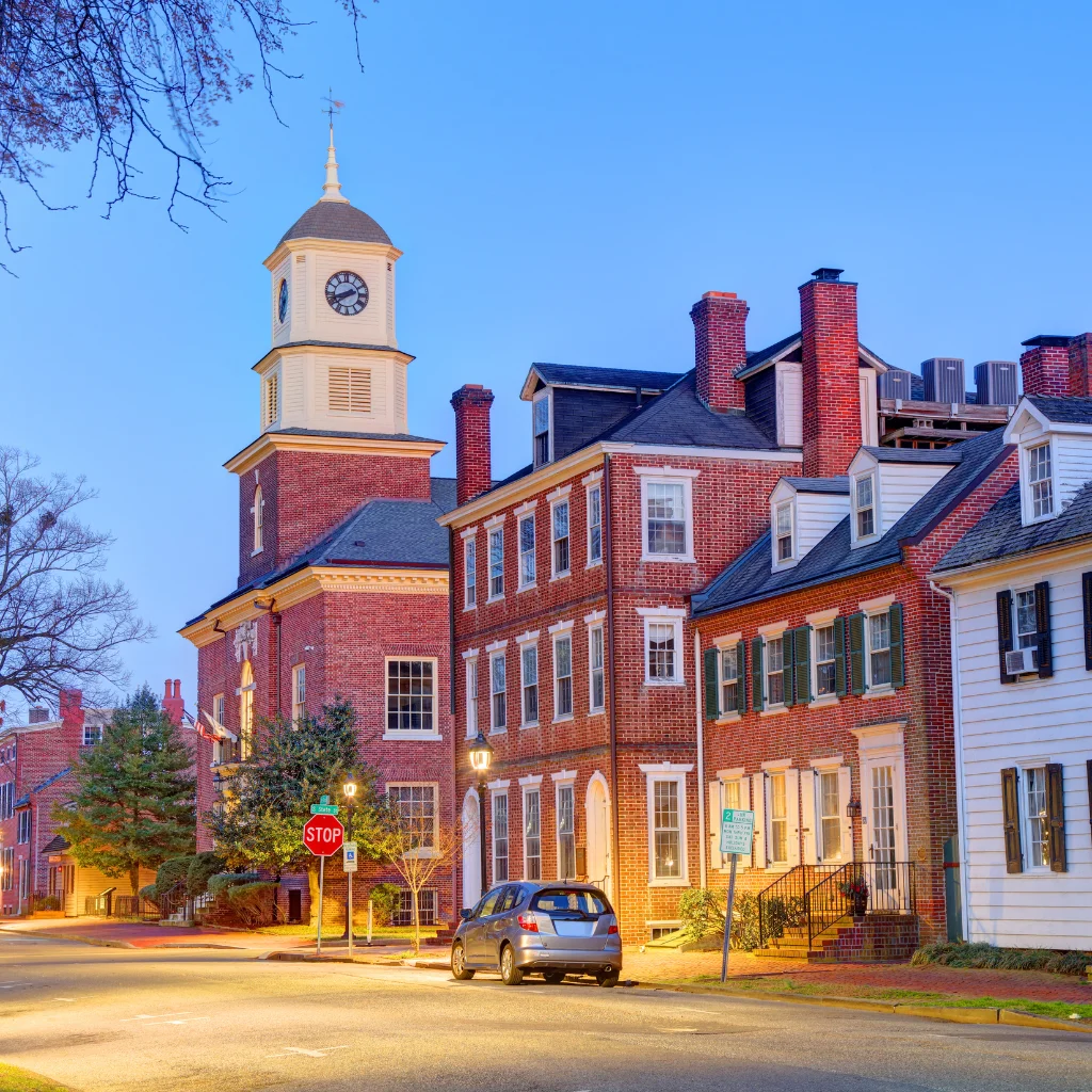 A charming evening view of a historic street in Delaware, featuring colonial-style red brick buildings and a clock tower. Warm streetlights illuminate the quiet neighborhood, adding to its timeless appeal. Transportvibe