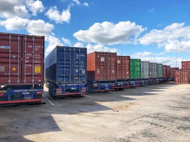 A row of colorful shipping containers on trailers parked in a logistics yard under a bright blue sky. Transportvibe.