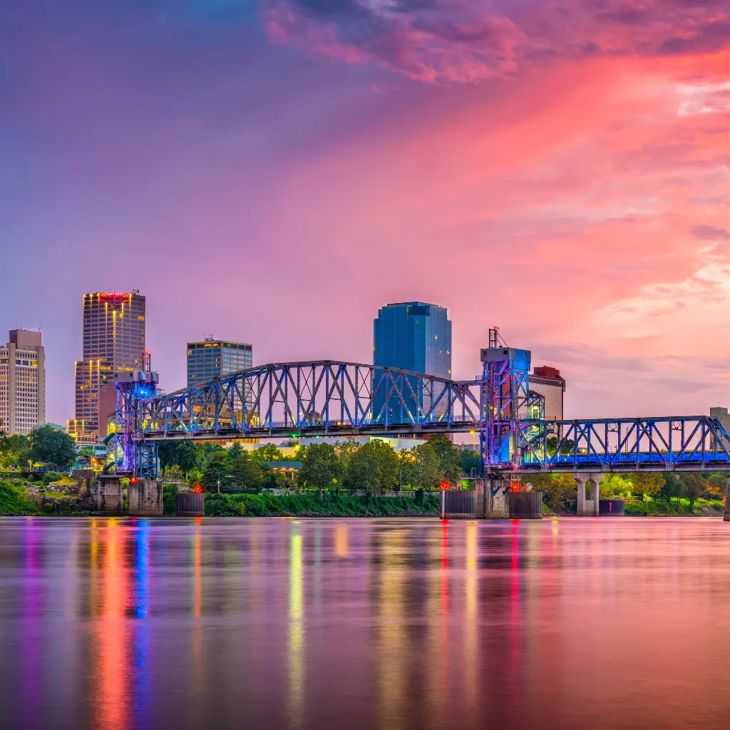 A vibrant view of the Little Rock, Arkansas skyline at sunset, featuring a colorful sky, illuminated bridge, and reflections on the Arkansas River. Transportvibe