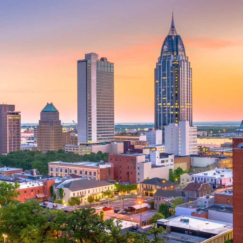 A stunning view of the Mobile, Alabama skyline at sunset, showcasing modern skyscrapers and historic buildings with a warm, colorful sky in the background. Transportvibe