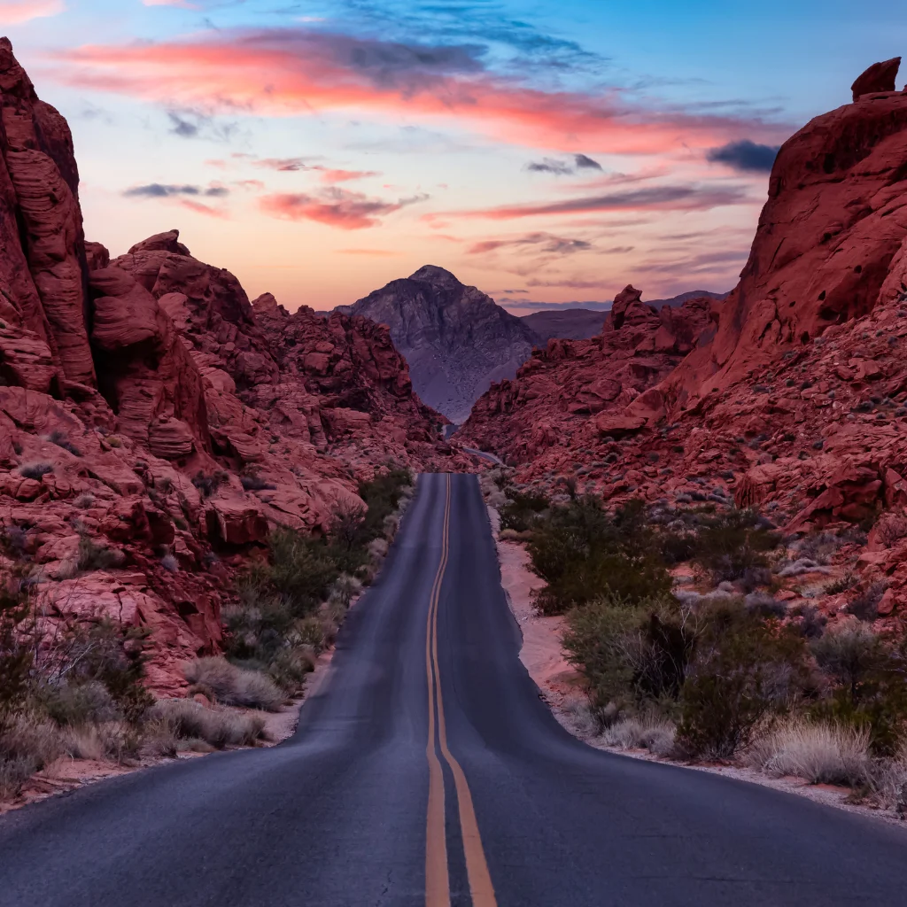 A scenic desert highway in Nevada surrounded by red rock formations at sunset. Transportvibe