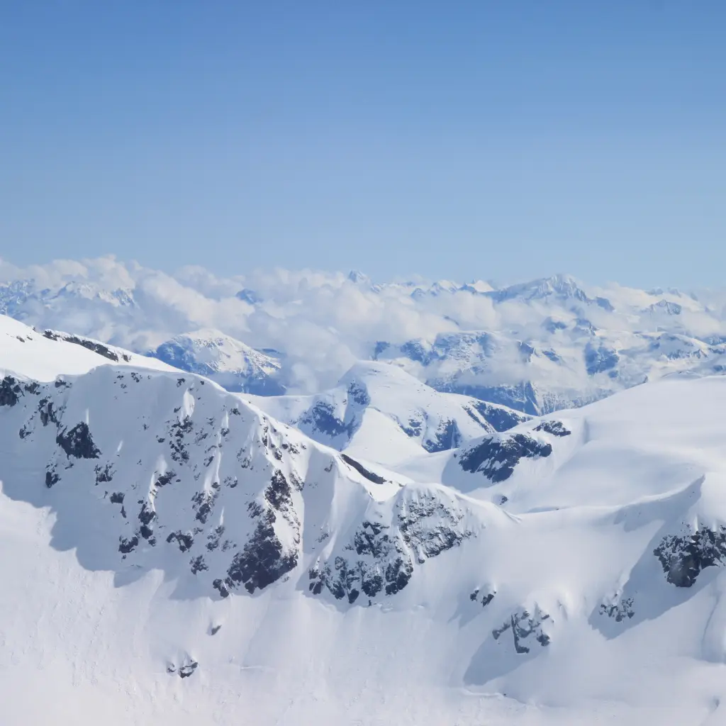 A breathtaking view of snow-covered mountain peaks in Alaska, with rugged slopes and a clear blue sky above. Transportvibe