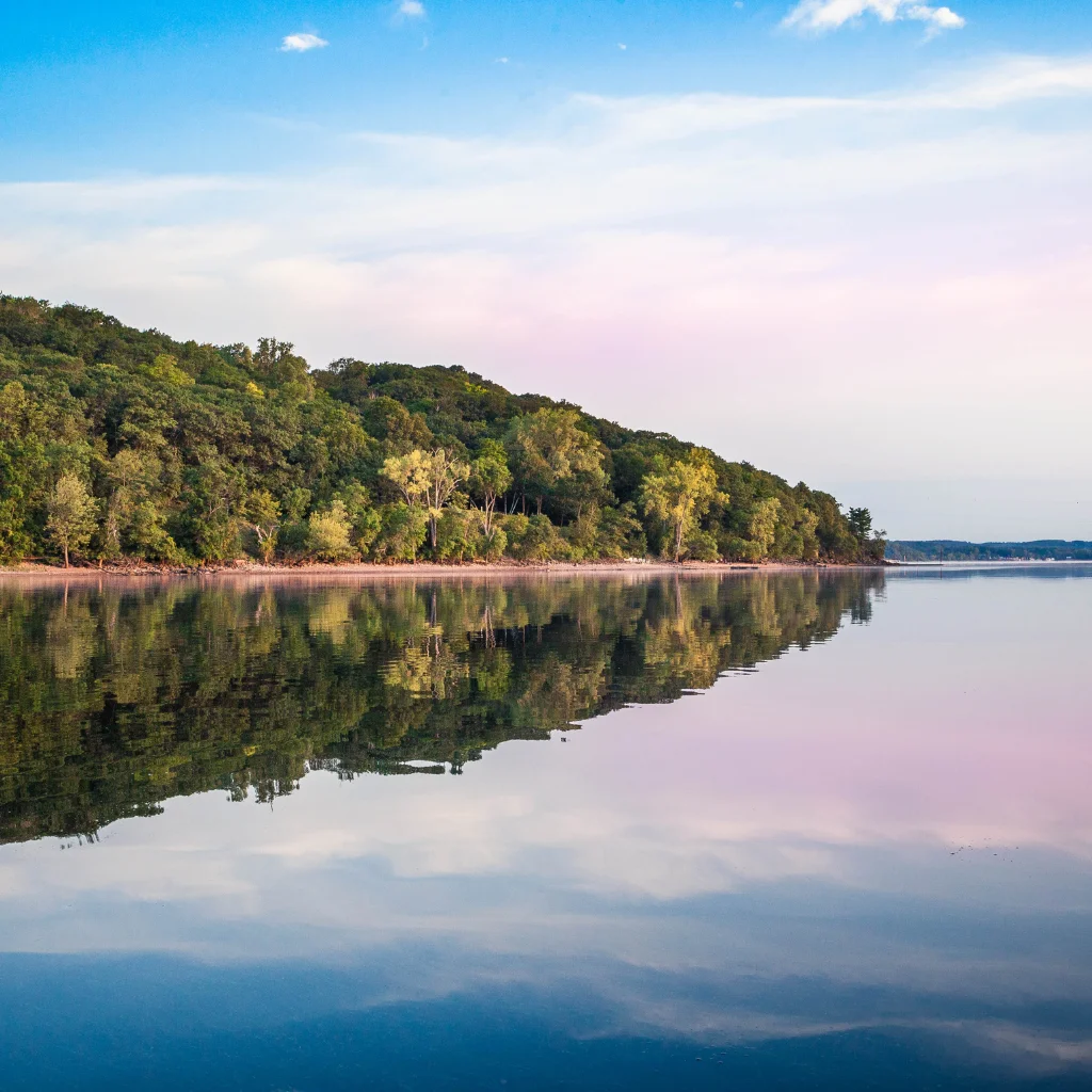 A tranquil lake in Wisconsin reflecting lush green trees and a pastel sky. Transportvibe